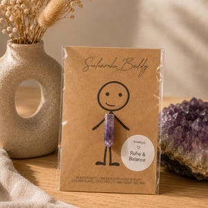 Può includere: Un piccolo ciondolo di cristallo di ametista viola su una carta marrone con un disegno a forma di omino stilizzato. La carta recita "Seelenruhe Buddy" e "Ruhe & Balance". Un vaso con fiori secchi e un gruppo di cristalli viola sono sullo sfondo.