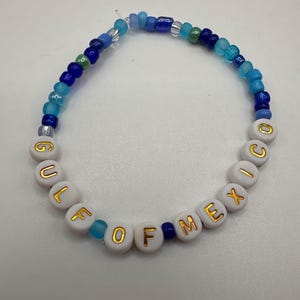 May include: A white beaded bracelet with the words "GULF OF MEXICO" spelled out in gold lettering. The beads are interspersed with blue and green glass beads.