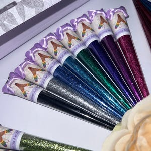 May include: A collection of henna art cones in various colors, including black, silver, blue, green, purple, and pink. Each cone is filled with glitter and has a decorative label with the text "Henna Art &" and a graphic. The cones are arranged in a fan shape.