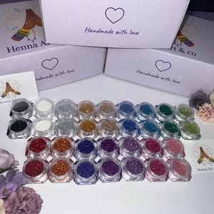 Könnte beinhalten: Eine Auswahl an Glitzer-Töpfen in verschiedenen Farben, darunter Schwarz, Weiß, Silber, Gold, Blau und Grün, in Reihen angeordnet. Das Bild zeigt auch Kartons mit dem Text "Henna Art & co" und "Handmade with love."