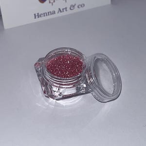 Könnte beinhalten: Ein kleines, klares, facettiertes Glas, gefüllt mit funkelndem, rötlich-rosa Glitzer. Der Deckel ist abgenommen und gibt den Inhalt frei. Im Hintergrund befindet sich eine Visitenkarte mit dem Text "Henna Art & co".