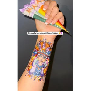 May include: A vibrant henna tattoo design on an arm, depicting a Native American woman with a feathered headdress. Several henna cones in shades of orange, green, and purple are held above the arm. Text on the image reads: Henna tattoo using coloured cones.