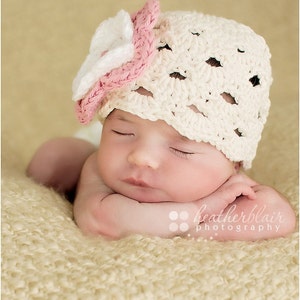 May include: A baby wearing a cream-coloured crocheted hat with a pink and white flower detail. The baby is lying on a soft, textured blanket, eyes closed. The image is a close-up shot.