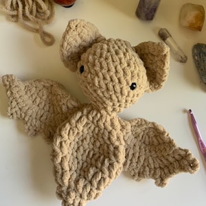 May include: A handmade, tan-colored crochet bat with black eyes and large, textured wings. The bat is lying on a white surface, with a ball of yarn, crystals, and a crochet hook nearby. The bat's ears are large and rounded.