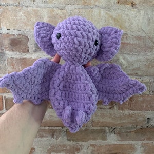 May include: A handmade, lavender-colored crochet bat plush toy. The bat has large, textured wings, big ears, and small black eyes. The plush is made of soft, chunky yarn, and is held against a brick wall background.