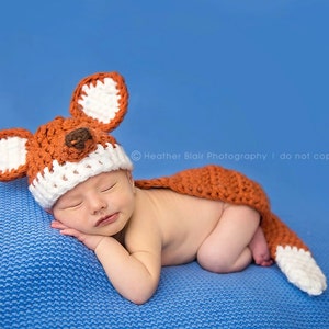 May include: A newborn baby wearing a crocheted fox hat and tail, sleeping on a blue blanket. The hat has white ears and a brown nose.