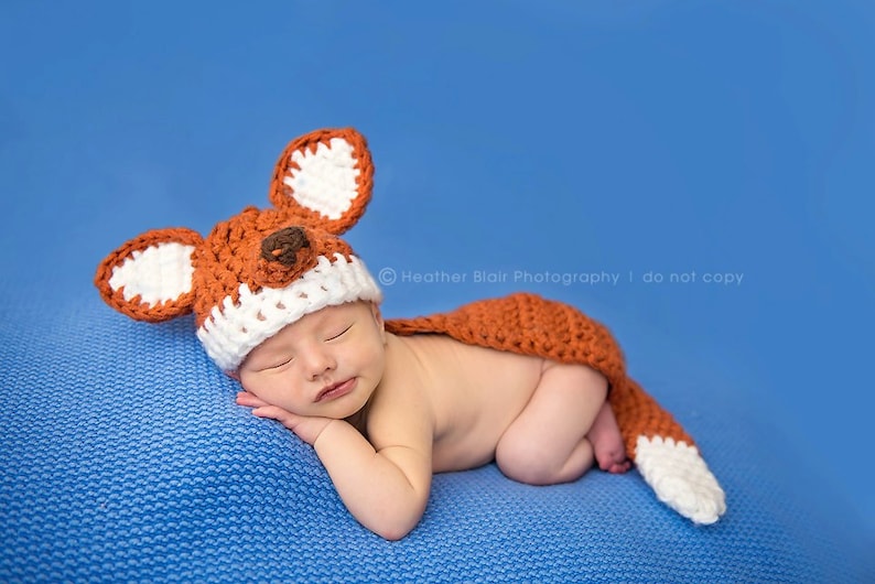 May include: A newborn baby wearing a crocheted fox hat and tail, sleeping on a blue blanket. The hat has white ears and a brown nose.