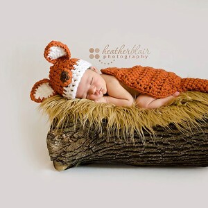 May include: A newborn baby wearing a crocheted fox costume, complete with a hat and tail, is sleeping peacefully on a log covered in faux fur. The baby is wearing a white and orange crocheted hat with fox ears and a matching orange crocheted tail.
