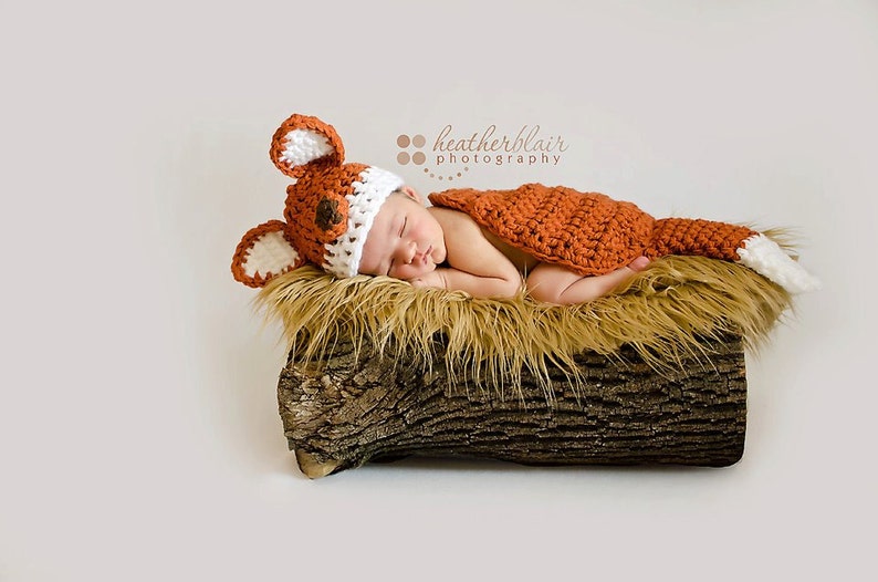 May include: A newborn baby wearing a crocheted fox costume, complete with a hat and tail, is sleeping peacefully on a log covered in faux fur. The baby is wearing a white and orange crocheted hat with fox ears and a matching orange crocheted tail.