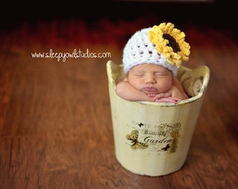 May include: A sleeping baby wearing a white crocheted hat with a yellow sunflower on top, resting in a white ceramic container with a butterfly garden design.