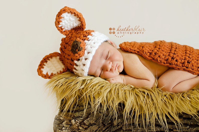 May include: A newborn baby wearing a crocheted fox costume, sleeping on a bed of faux fur. The costume is brown and white with a brown nose and ears.