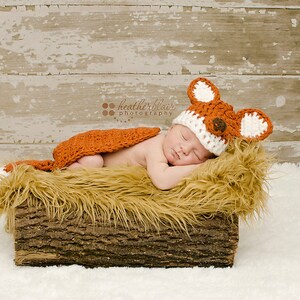 May include: A newborn baby wearing a crocheted fox hat and matching outfit is sleeping in a log-shaped bed filled with faux fur. The baby is wearing a white and orange crocheted fox hat with brown felt ears and a brown felt nose. The baby is wearing a matching orange crocheted fox outfit. The baby is sleeping on a bed of faux fur that is inside a log-shaped bed. The log-shaped bed is made of wood and has a rough texture.