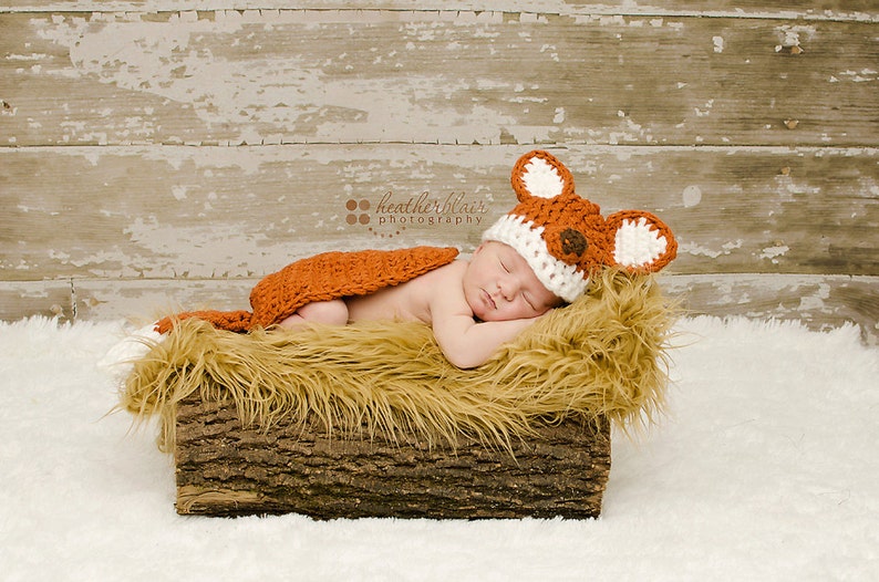 May include: A newborn baby wearing a crocheted fox hat and matching outfit is sleeping in a log-shaped bed filled with faux fur. The baby is wearing a white and orange crocheted fox hat with brown felt ears and a brown felt nose. The baby is wearing a matching orange crocheted fox outfit. The baby is sleeping on a bed of faux fur that is inside a log-shaped bed. The log-shaped bed is made of wood and has a rough texture.