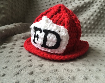 May include: A handmade red and white crocheted firefighter hat. The hat features a white patch with the letters "FD" in black. The hat is designed with a brim and a rounded top, resembling a miniature firefighter helmet.