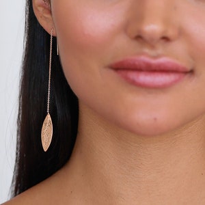 Puede incluir: Una mujer con el pelo largo y negro que lleva un top blanco sin mangas y pendientes de hoja de oro.