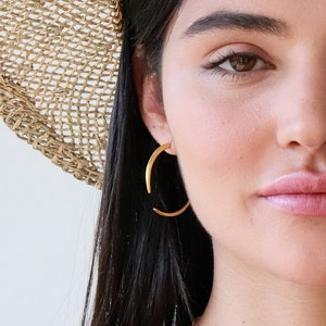 Peut inclure: Une femme portant un chapeau de paille, un débardeur noir et des bijoux en or. Elle porte un collier avec un pendentif en forme de feuille d'or et des boucles d'oreilles créoles en or.