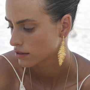 May include: A woman wearing a white dress with a gold necklace and earrings. She is also wearing a gold bracelet with a leaf design.