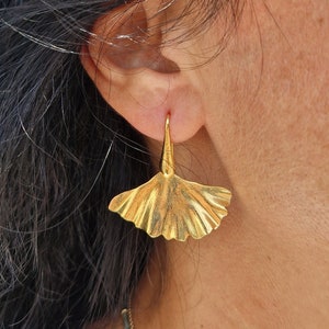 Op de afbeelding: Gouden ginkgo-blaadjes oorbellen met een delicaat ontwerp. De oorbellen zijn hangend en hebben een eenvoudige, elegante stijl.