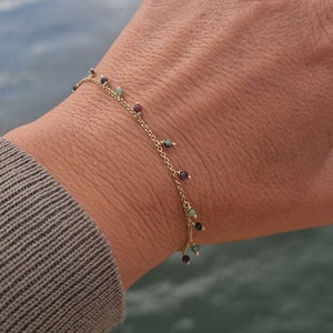 May include: A gold chain bracelet with small, colorful gemstones dangling from it. The bracelet is on a person's wrist.