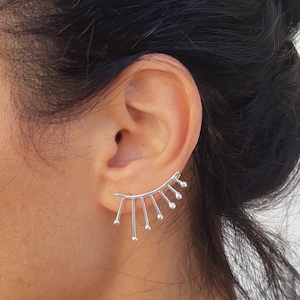 Może przedstawiać: Srebrny nausznik z motywem promieni słonecznych. Nausznik ma zakrzywiony pręt z pionowymi liniami i małymi, okrągłymi akcentami. Biżuteria jest noszona na uchu, prezentując swój nowoczesny i minimalistyczny styl.