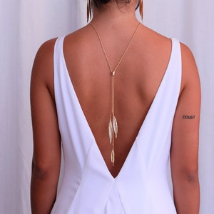 Può includere: Una donna indossa un abito bianco senza maniche con una profonda scollatura a V. Indossa una collana d'oro con un lungo ciondolo che le scende lungo la schiena. Il ciondolo è fatto di diverse piccole foglie dorate. La donna ha un tatuaggio sul braccio destro che dice "dubbio".