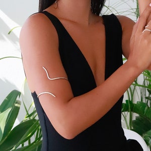 May include: A silver arm cuff with a curved design. The cuff is worn on the upper arm of a person wearing a black sleeveless top.