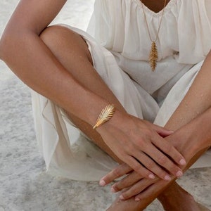 May include: A woman wearing a white dress sits with her legs crossed on a white sandy surface. She is wearing gold leaf earrings, a gold leaf necklace, and a gold leaf bracelet.