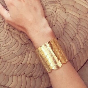以下が含まれることがあります： 鱗模様のテクスチャが施されたゴールドトーンのブレスレット。ブレスレットは手首に着用されています。