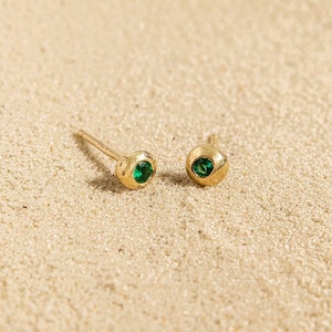 May include: Two gold stud earrings, each featuring a small, round, emerald-green gemstone. The earrings are set in textured gold and rest on a sandy surface, offering a simple yet refined appearance.