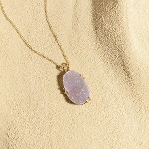 Puede incluir: Un delicado collar de cadena dorada con un gran colgante de forma ovalada hecho de una piedra de color púrpura claro, brillante y texturizada.