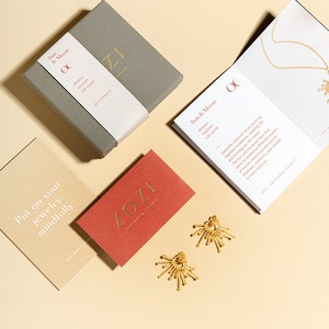 K&ouml;nnte beinhalten: Ein Paar goldene Ohrringe mit einem Sonnenstrahlen-Design, eine kleine Schachtel mit einem goldenen Logo und eine Karte mit dem Text "Sun & Moon Balance Intuition Life Cycles" in goldenen Lettern. Der Hintergrund ist hellbeige.