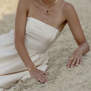 K&ouml;nnte beinhalten: Eine Frau in einem wei&szlig;en tr&auml;gerlosen Kleid mit einer goldenen Halskette und einem goldenen Ring. Die Halskette hat einen schwarzen Stein in der Mitte und der Ring hat ein Sternenmuster. Die Frau sitzt im Sand.