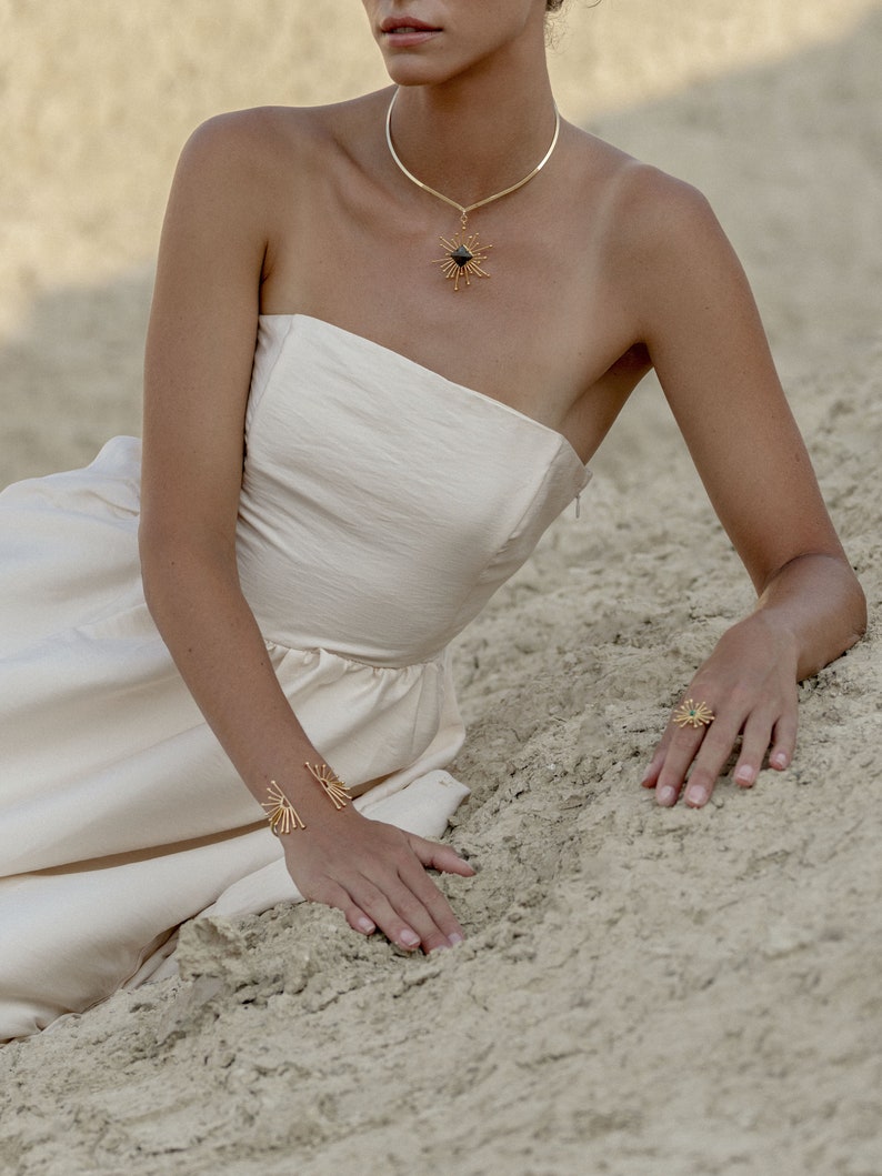 K&ouml;nnte beinhalten: Eine Frau in einem wei&szlig;en tr&auml;gerlosen Kleid mit einer goldenen Halskette und einem goldenen Ring. Die Halskette hat einen schwarzen Stein in der Mitte und der Ring hat ein Sternenmuster. Die Frau sitzt im Sand.