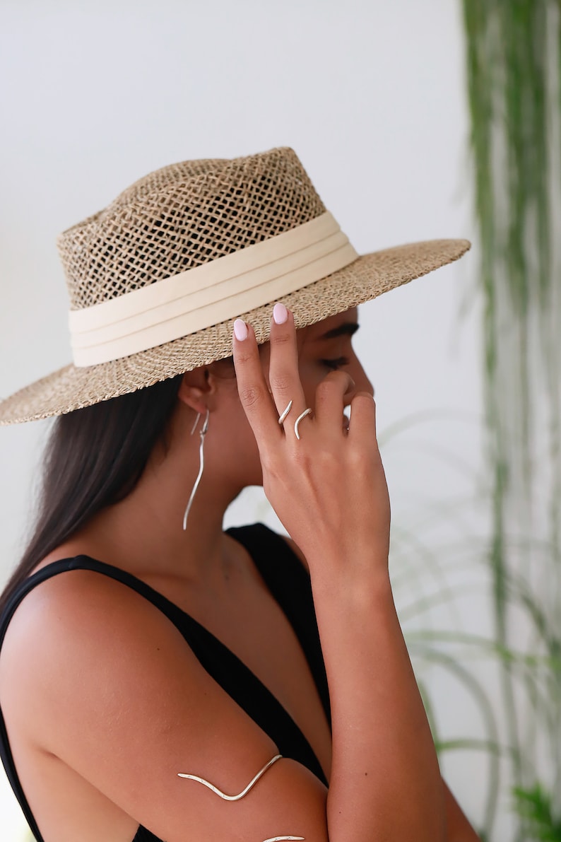 May include: A woman wearing a wide-brimmed straw hat with a white band, a black tank top, and silver jewelry. The hat is tilted down over her eyes.
