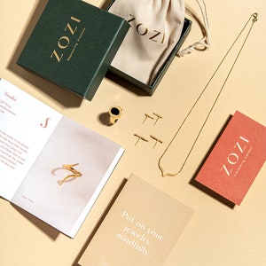 May include: A flat lay of gold jewelry, including a necklace, earrings, and a ring, displayed on a beige surface. The jewelry is packaged in a green box with the word "ZOZI" printed on it. There is also a small beige pouch with the word "ZOZI" printed on it. A card with the text "Put on your jewelry mindfully" is also included.