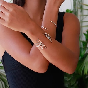 K&ouml;nnte beinhalten: Zwei silberne Armb&auml;nder am Handgelenk einer Person. Die Armb&auml;nder sind aus d&uuml;nnem Metall und haben ein Sonnenstrahlen-Design.