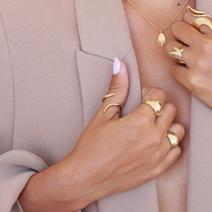 May include: A woman's hand wearing several gold rings, including a snake ring, a textured band, and a large, textured ring. The hand is holding a gold necklace with a hand charm.