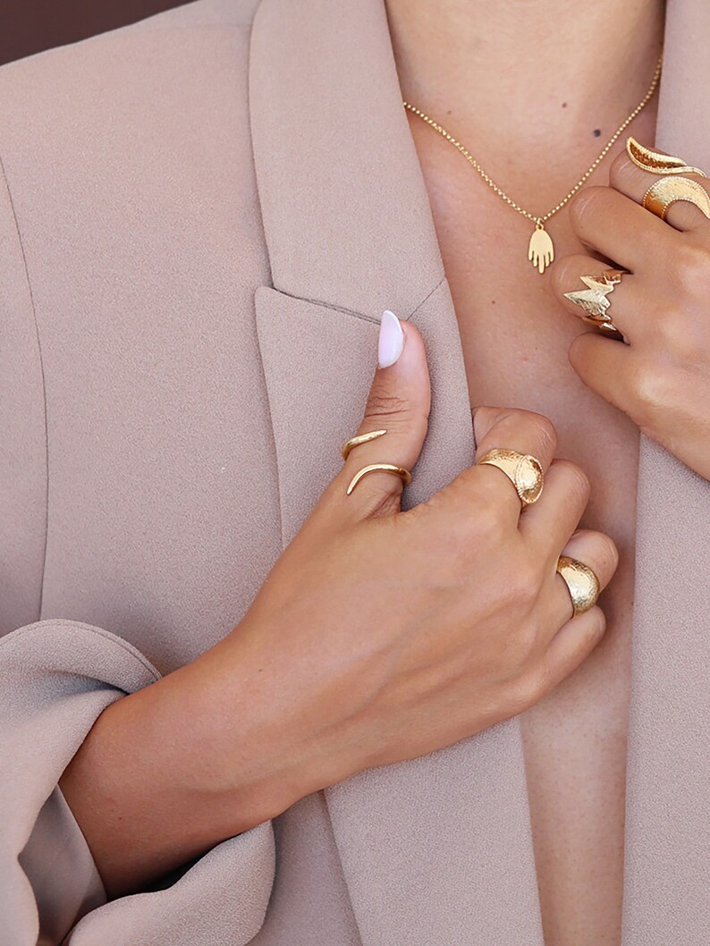 May include: A woman's hand wearing several gold rings, including a snake ring, a textured band, and a large, textured ring. The hand is holding a gold necklace with a hand charm.
