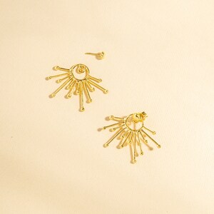 May include: A pair of gold earrings with a circular design and multiple radiating points. The earrings have a simple, modern style.