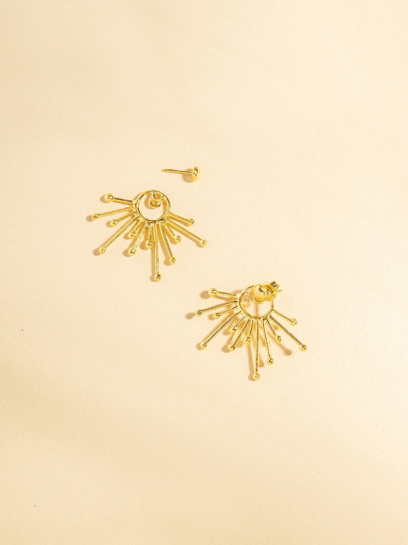 May include: A pair of gold earrings with a circular design and multiple radiating points. The earrings have a simple, modern style.