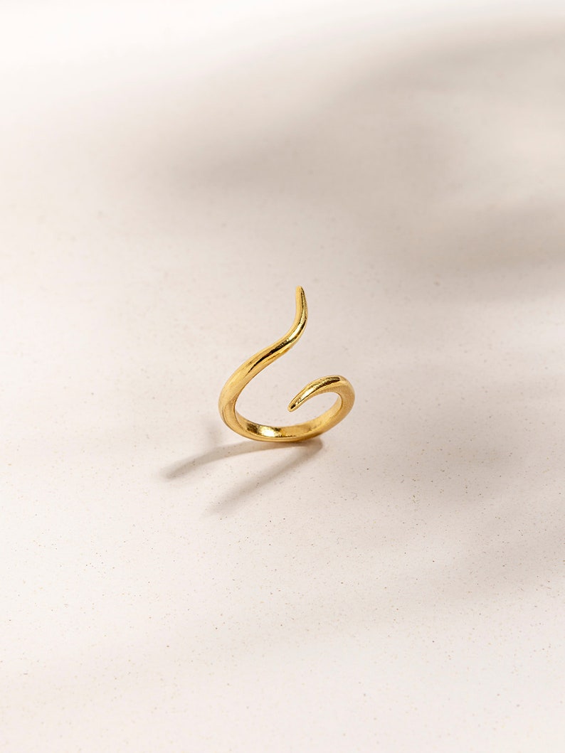 May include: A gold-toned ring with a unique, twisted design. The ring is open and has a pointed end.