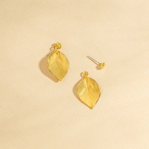 Puede incluir: Un par de pendientes de hoja de oro con un acabado texturizado. Los pendientes están sobre un fondo beige claro.