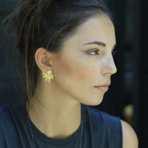 May include: A woman with dark brown hair wearing a dark gray sleeveless shirt and gold earrings. The earrings are a geometric design with multiple points.