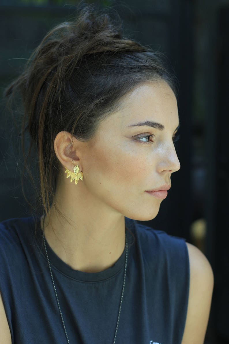May include: A woman with dark brown hair wearing a dark gray sleeveless shirt and gold earrings. The earrings are a geometric design with multiple points.