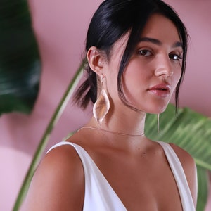Pode incluir: Uma mulher vestindo um top branco sem mangas com um decote em V profundo e brincos de folha de ouro. Ela tem cabelo escuro puxado para tr&aacute;s em um rabo de cavalo.