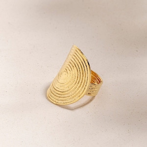 May include: A gold ring featuring a large, textured, half-circle design. The ring is crafted from metal and has a smooth, polished finish.