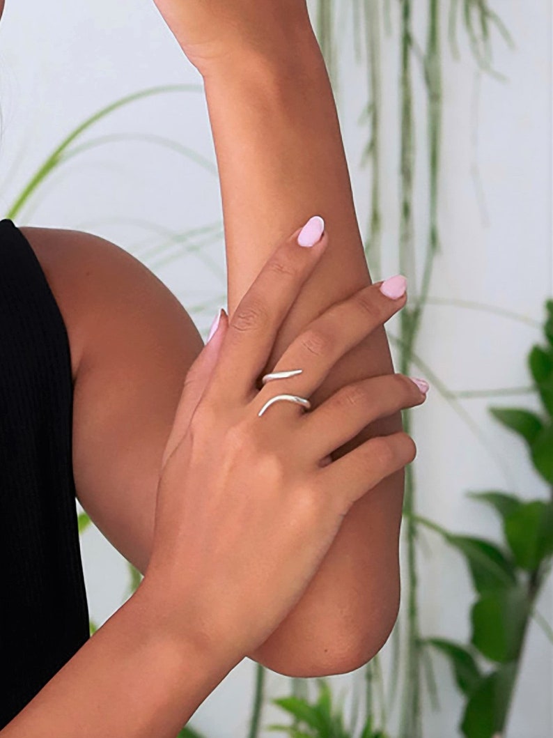 May include: A silver ring with a curved design. The ring is worn on the middle finger of a person's hand.
