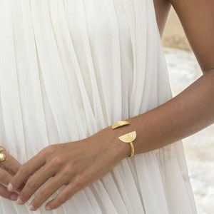 May include: A gold bracelet with two half-circle shapes, one with a textured surface and the other with concentric circles. The bracelet is worn on a person's wrist.