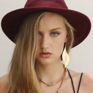 Peut inclure: Une femme aux longs cheveux blonds porte un chapeau fedora bordeaux et des boucles d'oreilles en feuille d'or. Elle porte également un collier superposé avec des cordons en cuir marron et des accents dorés.