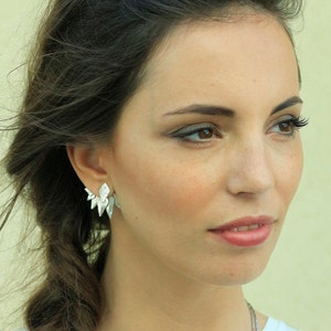 May include: A woman with long brown hair wearing a white shirt and a silver necklace. She is wearing silver earrings that are shaped like points.